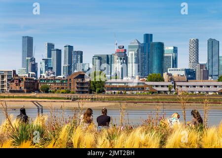 Menschen, die am Ufer des Greenwich mit Blick auf die Wolkenkratzer von Canary Wharf sitzen, London, Großbritannien Stockfoto