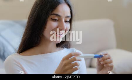 Porträt von freudig glücklich schwanger Frau Zukunft Mutter sitzt zu Hause hält Schwangerschaft positive Testergebnisse. Schöne arabische Latino junge Hausfrau Stockfoto