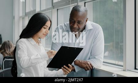 Verschiedene afrikanische und arabische Kollegen multirassische Menschen Geschäftspartner stehen in der Nähe des Fensters diskutieren Papier Grafiken strategische Pläne lesen Stockfoto