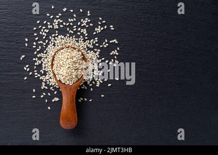 Rohe Sesamsamen in einem Holzlöffel auf schwarzem Schiefer-Hintergrund. Sesamum indicum für eine gesunde Ernährung und die Stärkung der Immunität Ernährung. Organische Benne-Körner. Stockfoto