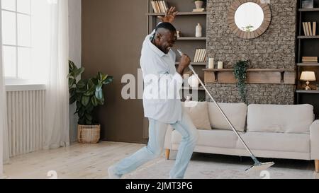 Aufgeregt künstlerischen Erwachsenen afrikanischen Mann Householder Bachelor aktiv Waschboden Reinigung Parkett mit Mopp zu Hause Wohnzimmer Umzug in Musik vortäuschend Stockfoto