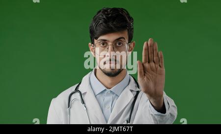 Junger männlicher Arzt Mann in medizinischer Maske isoliert in grünem Chroma-Schlüssel Studio zeigen Stopp-Schild mit unerschwinglicher Handbewegung sicheren Abstand stellt Handfläche nach vorne Stockfoto