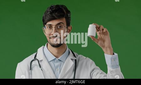 Indische männliche junge Arzt Berater in medizinischen Mantel stehen über grünem Hintergrund empfehlen bei der Kamera Flasche mit Schmerzmittel Pillen Vitamine Tabletten Stockfoto