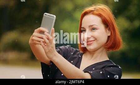 Portrait stilvolle schöne Mädchen kaukasisch attraktive rothaarige Frau blogger weibliches Modell steht draußen im Park hält Smartphone nimmt Selfie Foto auf Stockfoto