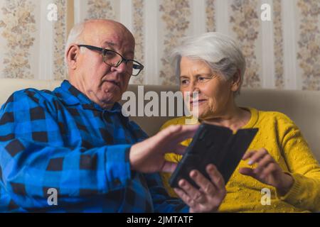 Kaukasisches Seniorenpaar kauft online mit dem Smartphone, während es auf einem Sofa im Wohnzimmer sitzt. Hochwertige Fotos Stockfoto