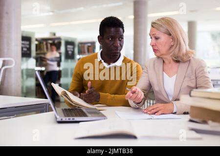 Freundliche weibliche Tutor hilft fleißigen afroamerikanischen Mann Vorbereitung für die Prüfung in der Bibliothek Stockfoto