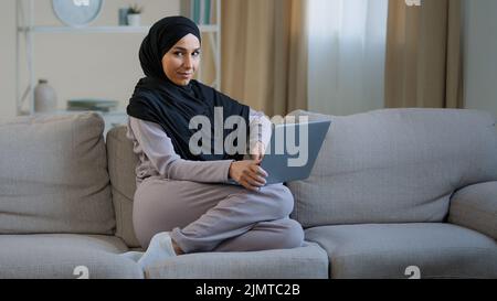 Attraktive junge muslimische Frau tragen Hijab sitzen auf gemütlichem Sofa im Wohnzimmer liebenswert energisch Millennial islamischen arabischen Mädchen hat Spaß im Wohnzimmer Stockfoto