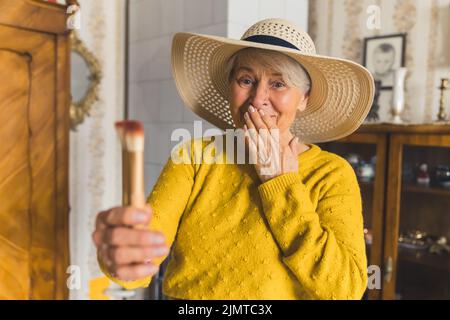 Lustige kaukasische ältere Frau, die zu Hause einen stylischen Strandhut trägt und Make-up-Bürsten in einer Hand hält und schockiert und verwirrt aussieht. Hochwertige Fotos Stockfoto