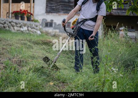 Junge kaukasische Mann Landwirt Gärtner auf dem Feld mit Saitenschneider Benzin Bürstenschneider Schnitt Unkrautgras arbeiten auf dem Bauernhof schneiden auf dem Feld i Stockfoto