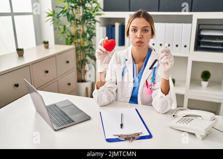 Junge blonde Frau trägt Arzt Uniform hält Blut und Herz puffing Wangen mit lustigen Gesicht. Mund mit Luft aufgeblasen, Luft fangen. Stockfoto