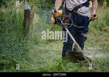 Junge kaukasische Mann Landwirt Gärtner auf dem Feld mit Saitenschneider Benzin Bürstenschneider Schnitt Unkrautgras arbeiten auf dem Bauernhof schneiden auf dem Feld i Stockfoto