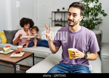 Hispanischer Vater einer interrassischen Familie, die lächelnd eine Tasse Kaffee trinkt und auf die Kamera schaut, die die Finger beim Siegeszeichen zeigt. Nummer zwei. Stockfoto