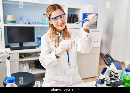 Junge blonde Frau trägt Wissenschaftler Uniform Gießen Flüssigkeit mit Pipette im Labor Stockfoto