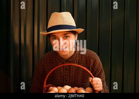 Viele Eier im Korb. Junge Frau im Hut hält ganze Korb von braunen Bio-Eier auf modernen grünen Hintergrund. Geflügelfarm. Umweltfreundliches Produkt. Landwirtschaft. Stockfoto