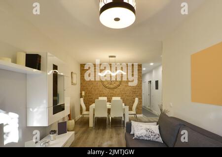 Esszimmer mit einem weißen Holztisch, weißen Lederpolsterstühlen und einer Backsteinwand kombiniert mit weißen Wänden in einem Wohnzimmer mit drei-er-Sitzmöbeln Stockfoto