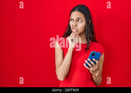 Junge brasilianische Frau mit Smartphone über rotem Hintergrund denken besorgt über eine Frage, besorgt und nervös mit der Hand auf dem Kinn Stockfoto