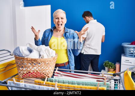 Hispanische Mutter und Sohn hängen Kleider an der Wäscheleine und feiern den Sieg mit glücklichen Lächeln und dem Siegerausdruck mit erhobenen Händen Stockfoto