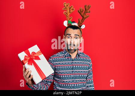 Junger hispanischer Mann mit Hirsch weihnachtshut mit Geschenk verängstigt und erstaunt mit offenem Mund für Überraschung, Unglauben Gesicht Stockfoto