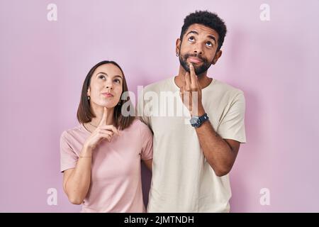 Junge hispanische Paar zusammen über rosa Hintergrund denken konzentriert über Zweifel mit Finger auf Kinn und Blick bis wundernd Stockfoto