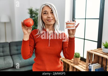 Grauhaarige Frau mittleren Alters, die selbstbewusst lächelt und roten Apfel und Prothese zu Hause hält Stockfoto
