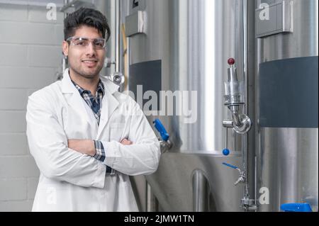 Junger, hübscher Arbeiter in weißem Laborkittel, lächelnd und zur Kamera schauend, stehend an Reihen von Stahlbraukbottichen und beim Überqueren Stockfoto