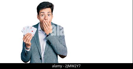 Junger hispanischer Geschäftsmann spielt Poker mit Karten, die den Mund mit der Hand bedecken, schockiert und Angst vor Fehlern. Überraschter Ausdruck Stockfoto