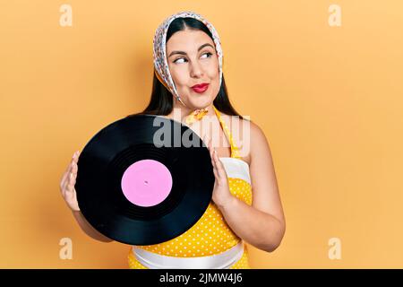 Junge hispanische Frau im Pin-up-Stil mit Vinyl-Scheibe, die lächelnd zur Seite schaut und denkend wegstarrt. Stockfoto