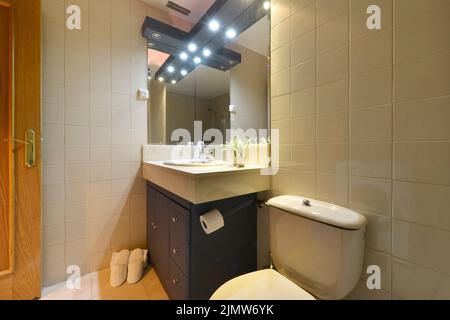 Badezimmer mit dunklen Holzschränken, rahmenlosen Spiegeln an beiden Seiten und weißen Hausschuhen Stockfoto