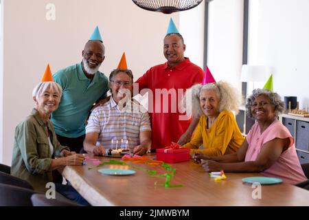 Porträt von fröhlichen, multirassischen Senioren, die Partyhüte tragen, während sie den Geburtstag eines Freundes feiern Stockfoto