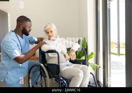 afroamerikanischer Gesundheitshelfer unterstützt kaukasische ältere Frau, die auf dem Rollstuhl sitzt Stockfoto