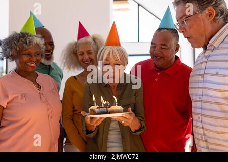 Glückliche multirassische Freunde tragen Partyhüte Blick auf ältere Frau weht Geburtstagskerzen Stockfoto