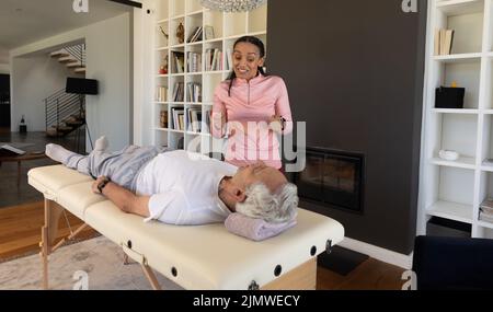 Biraziale Physiotherapeutin, die gestikuliert, während sie den kaukasischen älteren Mann führt, der zu Hause auf dem Tisch liegt Stockfoto