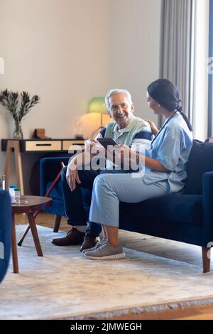 Biraziale Physiotherapeutin mit digitalem Tablet, während sie mit kaukasischem älteren Mann auf dem Sofa spricht Stockfoto