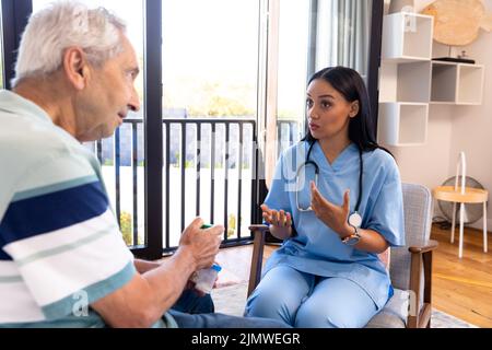 Biracial Physiotherapeutin im Gespräch mit kaukasischen älteren Mann, während auf Stuhl zu Hause sitzen Stockfoto