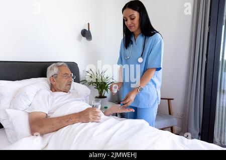 Biraziale Physiotherapeutin, die Medikamente an kaukasischen älteren Mann einnahm, der zu Hause auf dem Bett liegt Stockfoto