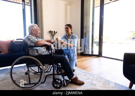 Biracial weibliche Physiotherapeutin reden und halten ältere Mann die Hand sitzt auf Rollstuhl zu Hause Stockfoto