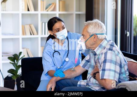 Biraziale Ärztin untersucht kaukasischen älteren Mann mit Sauerstoffmaske sitzen auf dem Sofa zu Hause Stockfoto