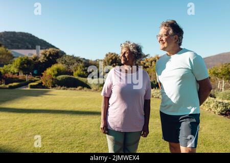 Multirassische ältere Freunde blicken weg, während sie auf grasbewachsenem Land gegen den klaren Himmel im Hof stehen Stockfoto