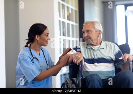 Lächelnde biraziale Physiotherapeutin im Gespräch mit kaukasischem Senioren, der auf dem Rollstuhl sitzt Stockfoto