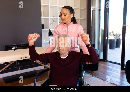 Biraziale Physiotherapeutin, die kaukasischen älteren Mann beim Heben von Hanteln im Rollstuhl unterstützt Stockfoto