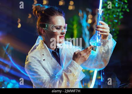 Moderne Wissenschaftlerin im Labormantel mit Kolben und futuristischer Brille in der virtuellen Realität. Stockfoto