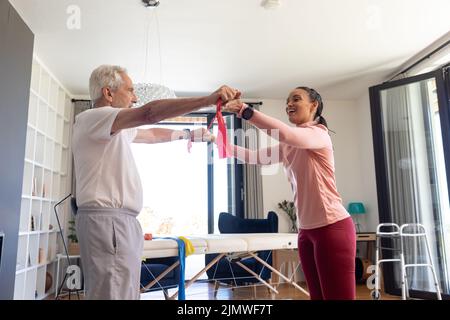 Biraziale Physiotherapeutin mit kaukasischem Senioren, der zu Hause das Widerstandsband streckt Stockfoto