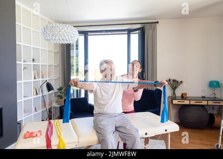 Biraziale Physiotherapeutin unterstützt kaukasischen älteren Mann beim Dehnen des Widerstandsbandes auf dem Tisch Stockfoto