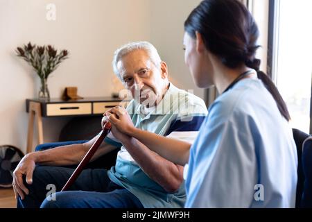 Biraziale Physiotherapeutin tröstet kaukasischen älteren Mann mit Gehstock auf dem Sofa sitzen Stockfoto