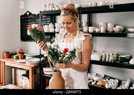 Dekorieren meines Ateliers. Eine attraktive reife Frau, die allein steht und in ihrer Töpferwerkstatt Blumen in eine handgemachte Vase setzt. Stockfoto