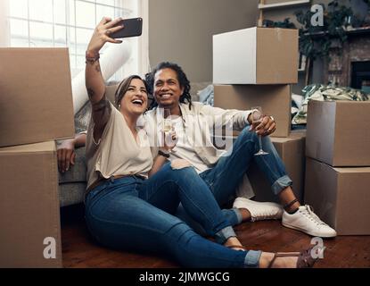 Wir zeigen unseren Freunden, was vor sich ging. Ein junges Paar, das ein Selfie macht, während es beschäftigt ist, in ihr neues Zuhause zu ziehen. Stockfoto