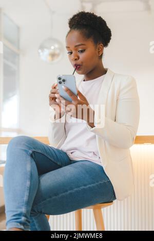 Porträt einer afroamerikanischen Frau, die ihre Freunde per SMS auf das Smartphone schreibt, in einem Café sitzt und ein Getränk trinkt. Internet-Technologie. Schönheit Stockfoto