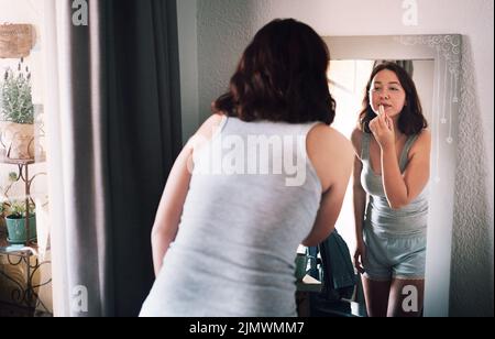Mal sehen, wie meine Lippen darin aussehen. Eine attraktive junge Frau, die Lippenstift aufbringt, während sie zu Hause vor dem Spiegel in ihrem Schlafzimmer steht. Stockfoto