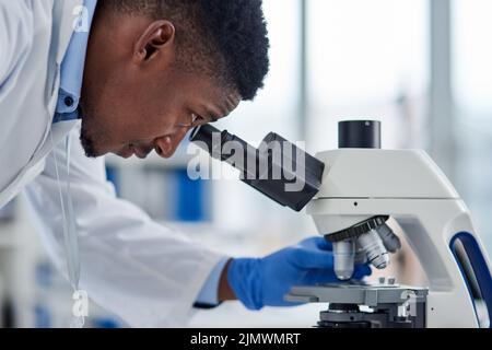 Schauen wir uns das einmal an. Ein fokussierter junger Wissenschaftler, der tagsüber durch ein Mikroskop in einem Labor Testproben ansieht. Stockfoto