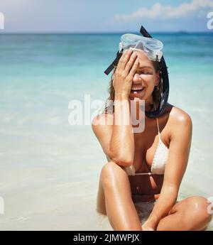 Sie kann gar nicht genug vom Wasser bekommen. Eine attraktive junge Frau in Tauchausrüstung lacht, während sie am Strand sitzt. Stockfoto